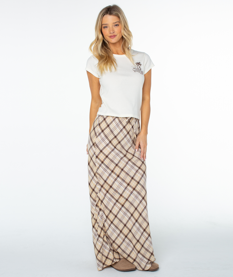 Woman standing against a white background in a fitted Kinda Salty 90S Baby Tee by Roxy with a small chest graphic, paired with a long beige and brown plaid skirt and tan shoes.