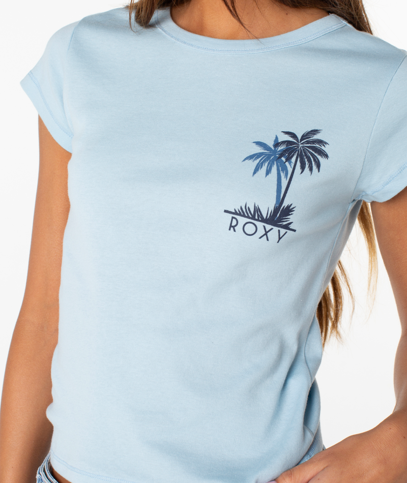 A person wears the Palm Geo 90S Baby Tee by Roxy—a light blue fitted tee with a small palm tree graphic and ROXY on the left chest. The photo shows only their upper torso, face not visible.