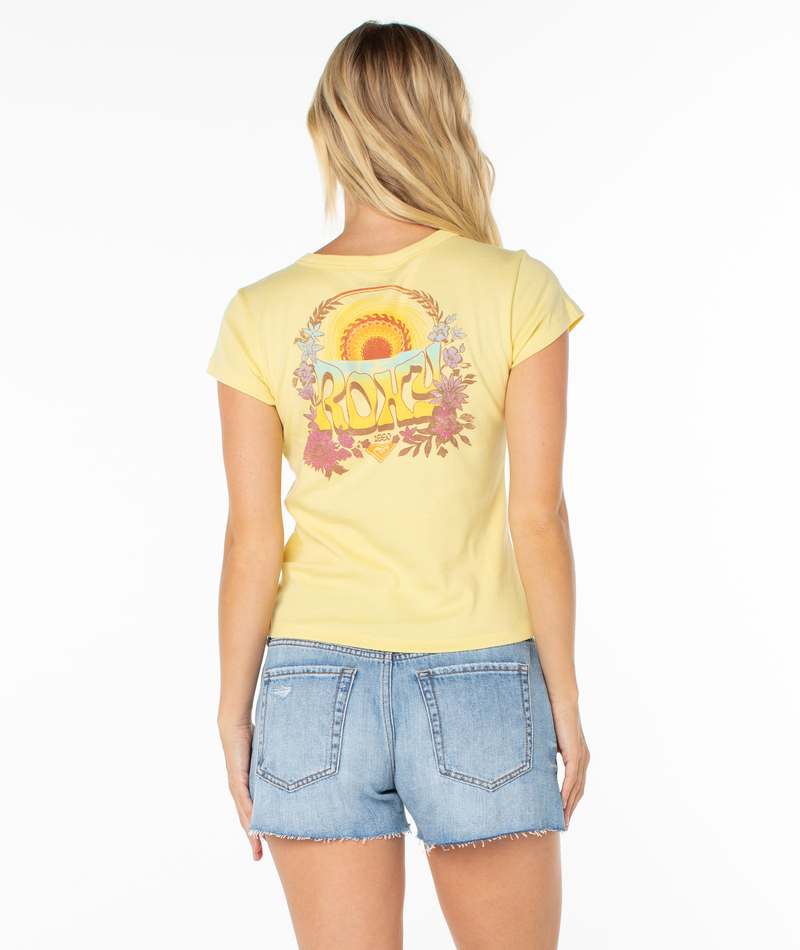 A woman with blonde hair wears the ROXY Tribal Sunset 90s Baby Tee, a yellow retro-inspired top with a colorful graphic, paired with light blue denim shorts, facing away and radiating beachy vibes against a white background.