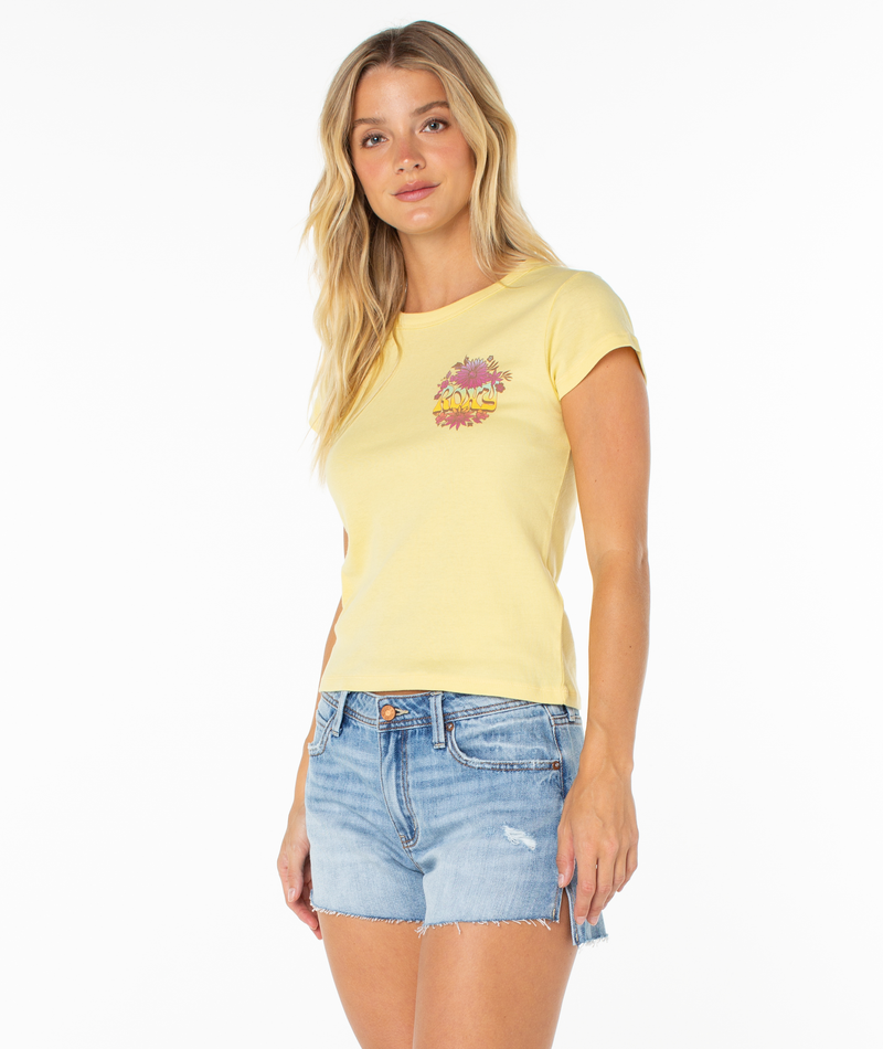 A woman with long blonde hair wears the Roxy Tribal Sunset 90S Baby Tee—a retro-inspired yellow t-shirt with a colorful chest graphic—paired with light blue denim shorts, standing against a plain white background.