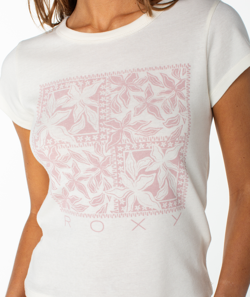 A woman wearing the Roxy Flower Tile 90S Baby Tee—a fitted white top featuring a light pink floral square pattern and “ROXY” printed at the hem, perfect for adding a retro-inspired touch to your wardrobe.
