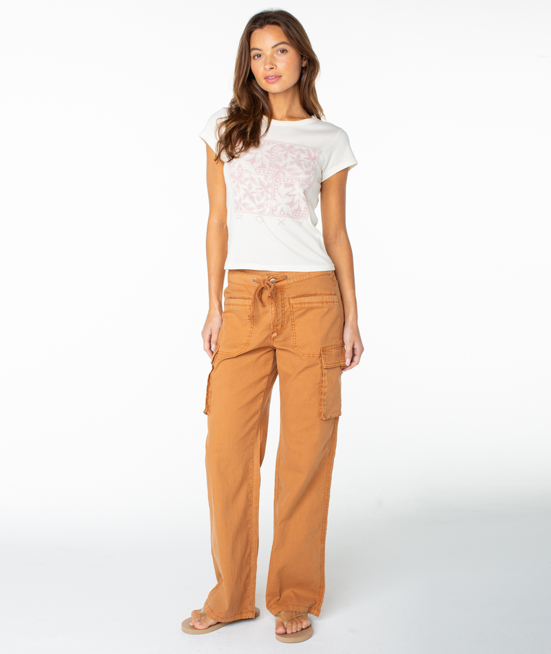 A woman faces forward against a plain white background, wearing the Roxy Flower Tile 90S Baby Tee paired with orange cargo pants and sandals. She has long brown hair and a neutral expression.