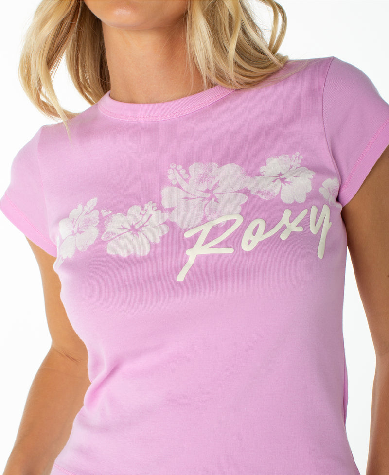 A person wearing the Roxy Floating Hibiscus 90S Baby Tee, a light pink, retro-inspired top featuring white hibiscus flowers and “Roxy” printed across the chest.
