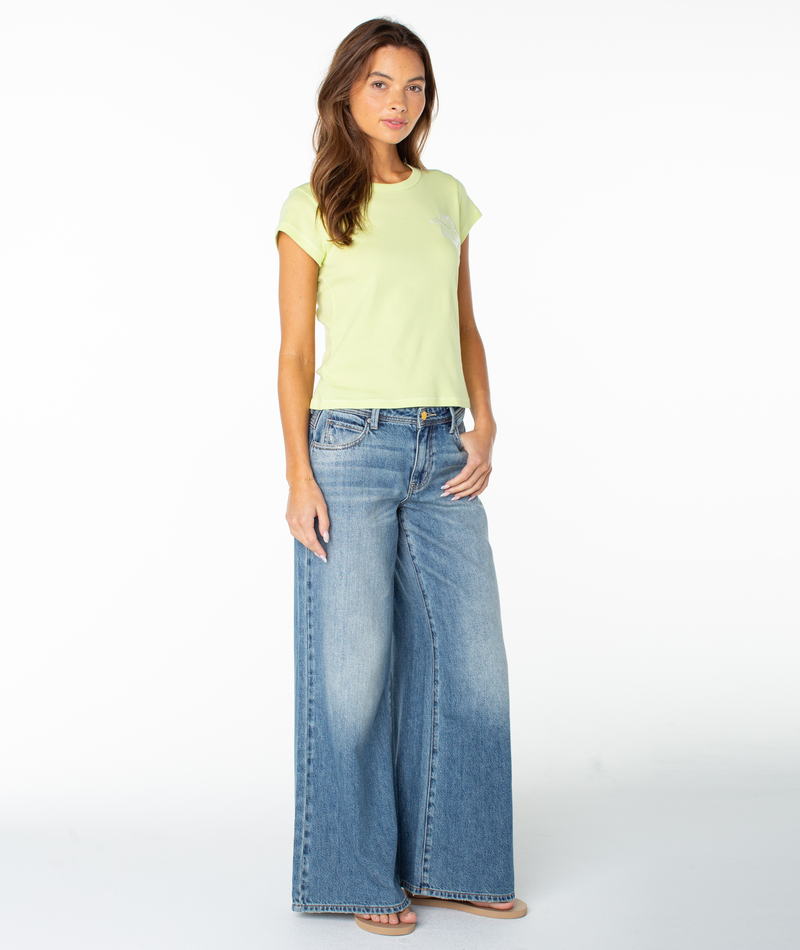 A woman with long brown hair and a neutral expression wears the Roxy Surf Day Embroidered 90S Baby Tee, a light yellow fitted t-shirt, paired with wide-leg blue jeans and sandals against a plain white background.