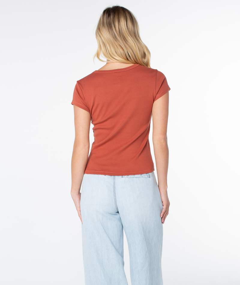 A woman with long blonde hair stands facing away, wearing the Roxy Shell Band 90S Baby Tee in a retro-inspired rust color and light blue jeans against a plain white background.