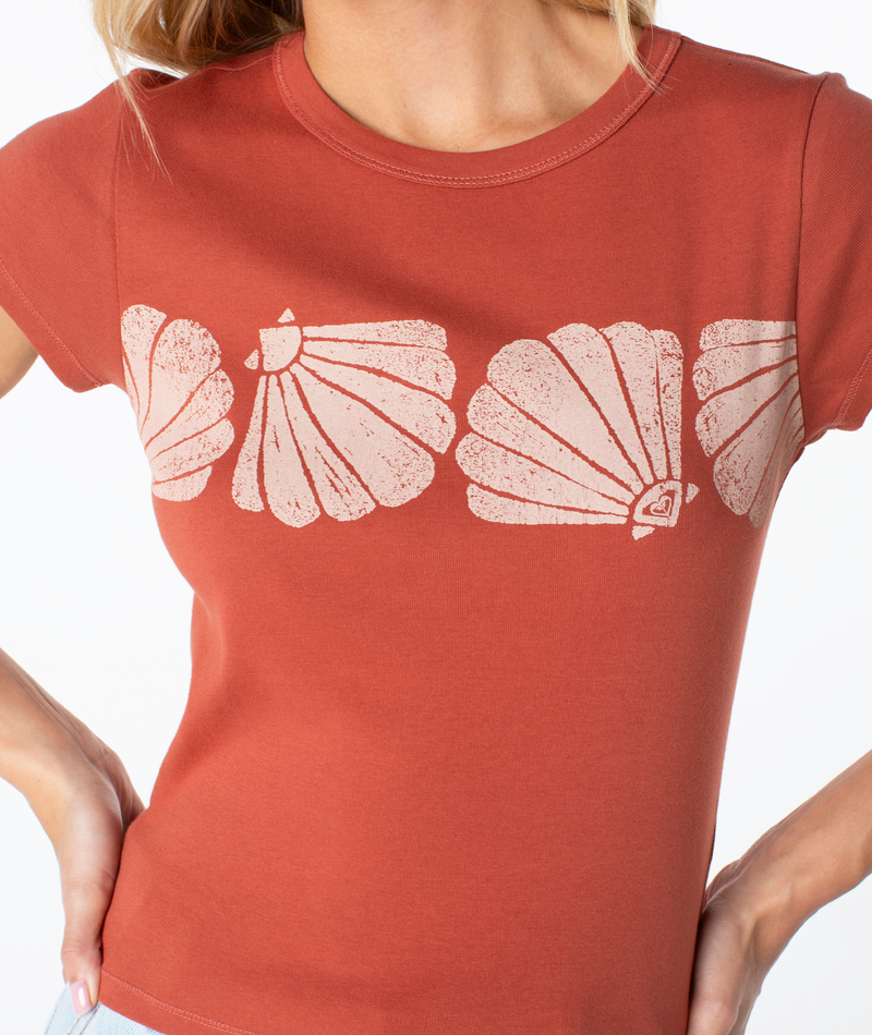 A person wearing the Roxy Shell Band 90S Baby Tee—a rust-colored, retro-inspired top featuring white shell band designs across the chest—stands with hands on hips.