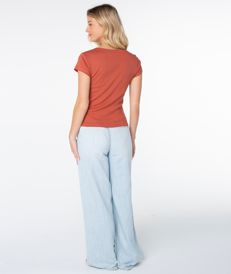 A woman stands facing away, wearing the Roxy Shell Band 90S Baby Tee in a rust shade and light blue wide-leg pants. The background is white, and she smiles softly with her head turned to the side.