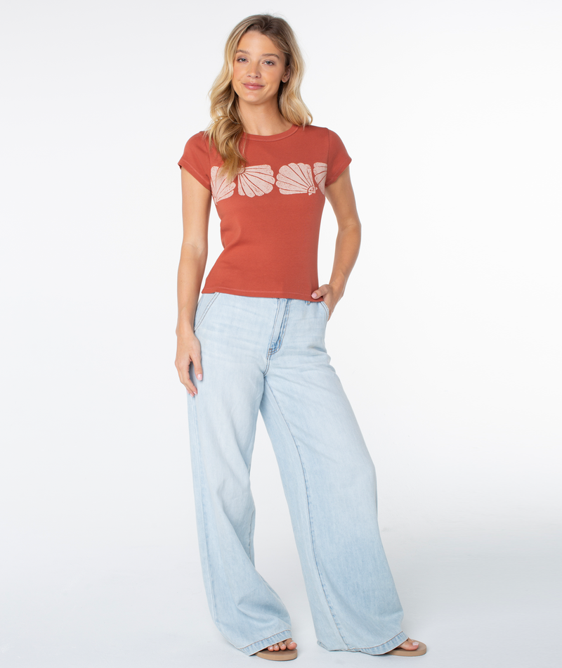 A woman stands against a white background wearing the Roxy Shell Band 90S Baby Tee, featuring shell designs across the chest, paired with light blue wide-leg jeans. She has long blonde hair and poses with one hand by her side and the other on her hip, channeling a subtly retro-inspired vibe.