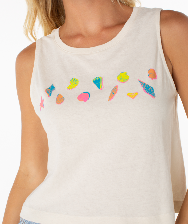 A woman in the Roxy Shell Risograph Tank—a sleeveless white top with vibrant seashell prints—exudes effortless beachy chic.