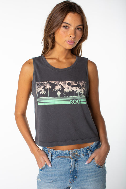 A woman with long brown hair wears a sleeveless dark gray Roxy Windy Palm Tank featuring a palm tree graphic and pairs it with light blue denim jeans, creating a cool beachy vibe against a plain white background.