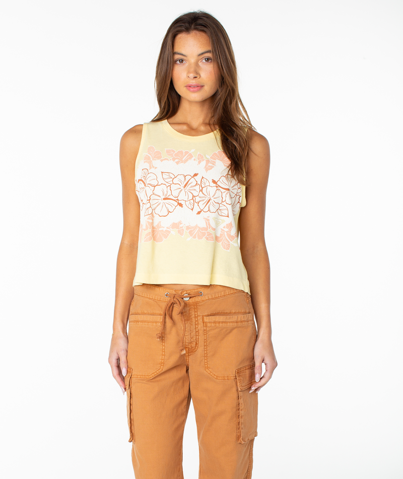 A woman with long brown hair wears the Roxy Hibiscus Lei Wildflower Tank in Golden Haze Hibiscus Lei, featuring a yellow sleeveless design with a pink floral pattern, paired with light brown cargo pants against a white background for an effortless tomboy style.