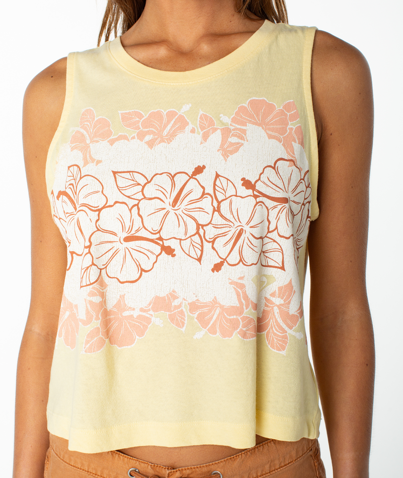 A woman wears the Roxy Hibiscus Lei Wildflower Tank, a sleeveless yellow crop top adorned with a bold peach and white hibiscus print across the chest and upper waist, capturing breezy, beachy chic style perfect for carefree sunny days.