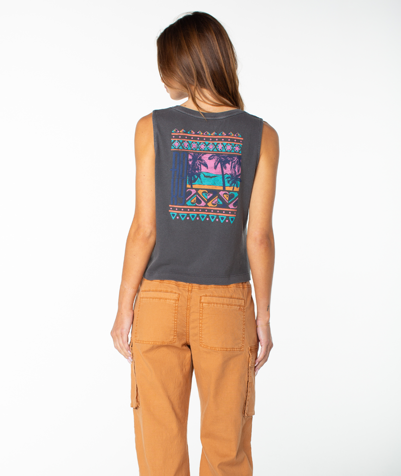 A woman with long brown hair stands facing away from the camera, wearing the Roxy Oceanview Wildflower Tank in Phantom Oceanview—a dark gray, sleeveless cropped top featuring a colorful tropical sunset graphic and geometric patterns that capture an effortless beachy chic vibe—paired with tan cargo pants against a plain white background.