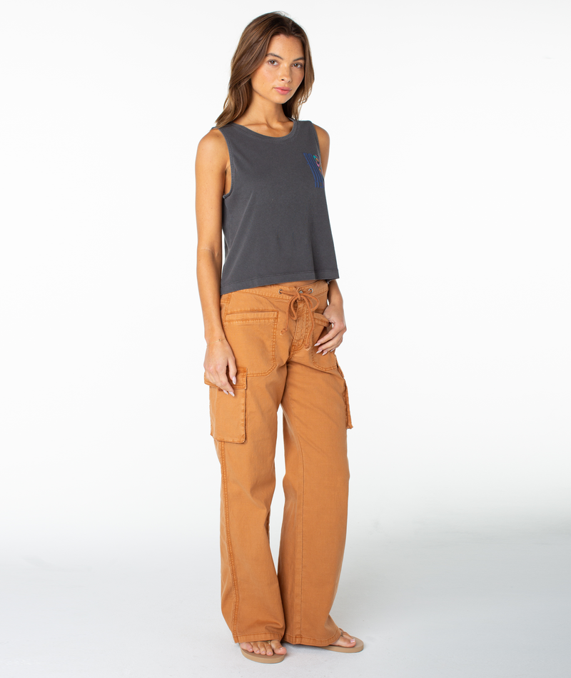 A woman stands in a studio wearing a sleeveless dark gray Roxy Oceanview Wildflower Tank and loose, high-waisted orange cargo pants with large pockets, paired with sandals. She exudes tomboy style as she poses with one arm relaxed by her side.