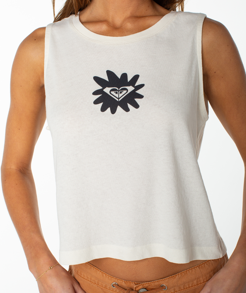 A woman wearing the Roxy Palm Poster Wildflower Tank, a sleeveless white top featuring a black flower and heart logo at the center, paired with relaxed light brown pants in a tomboy style, stands against a white background.