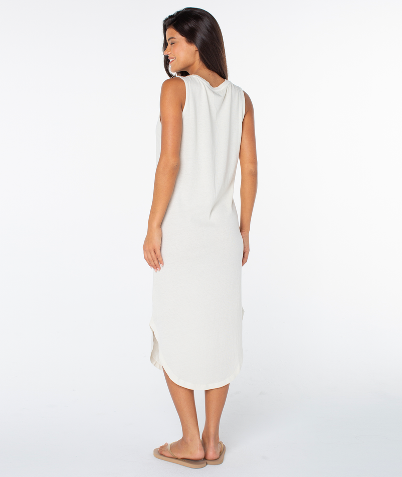 A woman stands facing away, wearing the Roxy Made In The Sun Wildflower Midi Dress and beige sandals, her long dark hair flowing down her back with her head slightly turned against a plain white background.