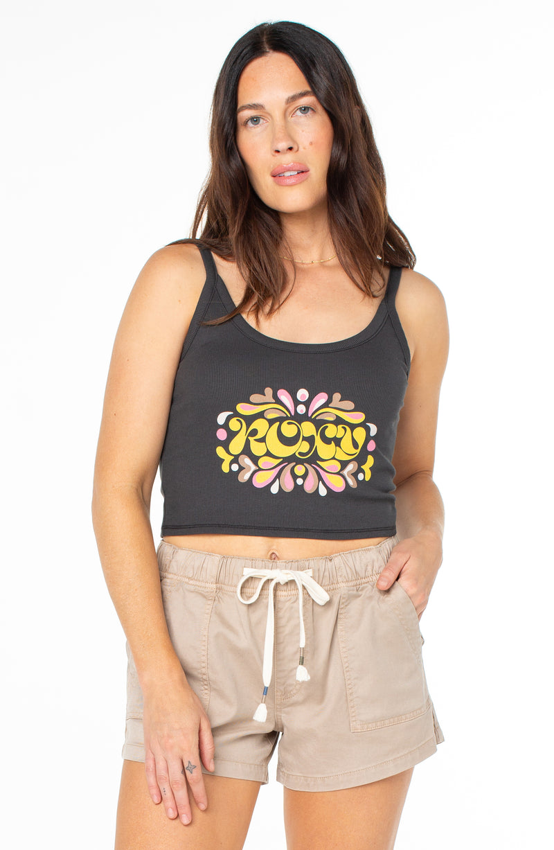 A woman wearing a Roxy Epic Vibe Wanderer Tank in Phantom Epic Vibe, featuring a yellow and pink floral-inspired graphic, with tan drawstring shorts.