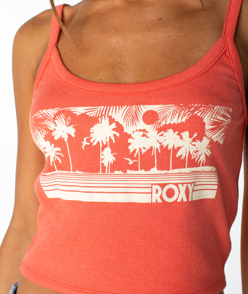 A woman wears the Roxy Windy Palm Wanderer Tank, a coral cropped top featuring a white palm tree, sun, and ROXY graphic on the front, made from a soft cotton blend for all-day comfort.