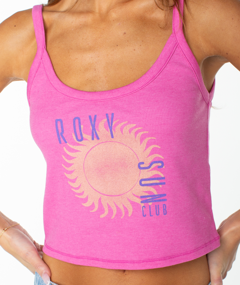 A person wears the Roxy Sun Club Wanderer Tank, a pink cotton blend top featuring "ROXY SUN CLUB" and a yellow sun graphic on the front. Only their torso and part of their arms are shown.