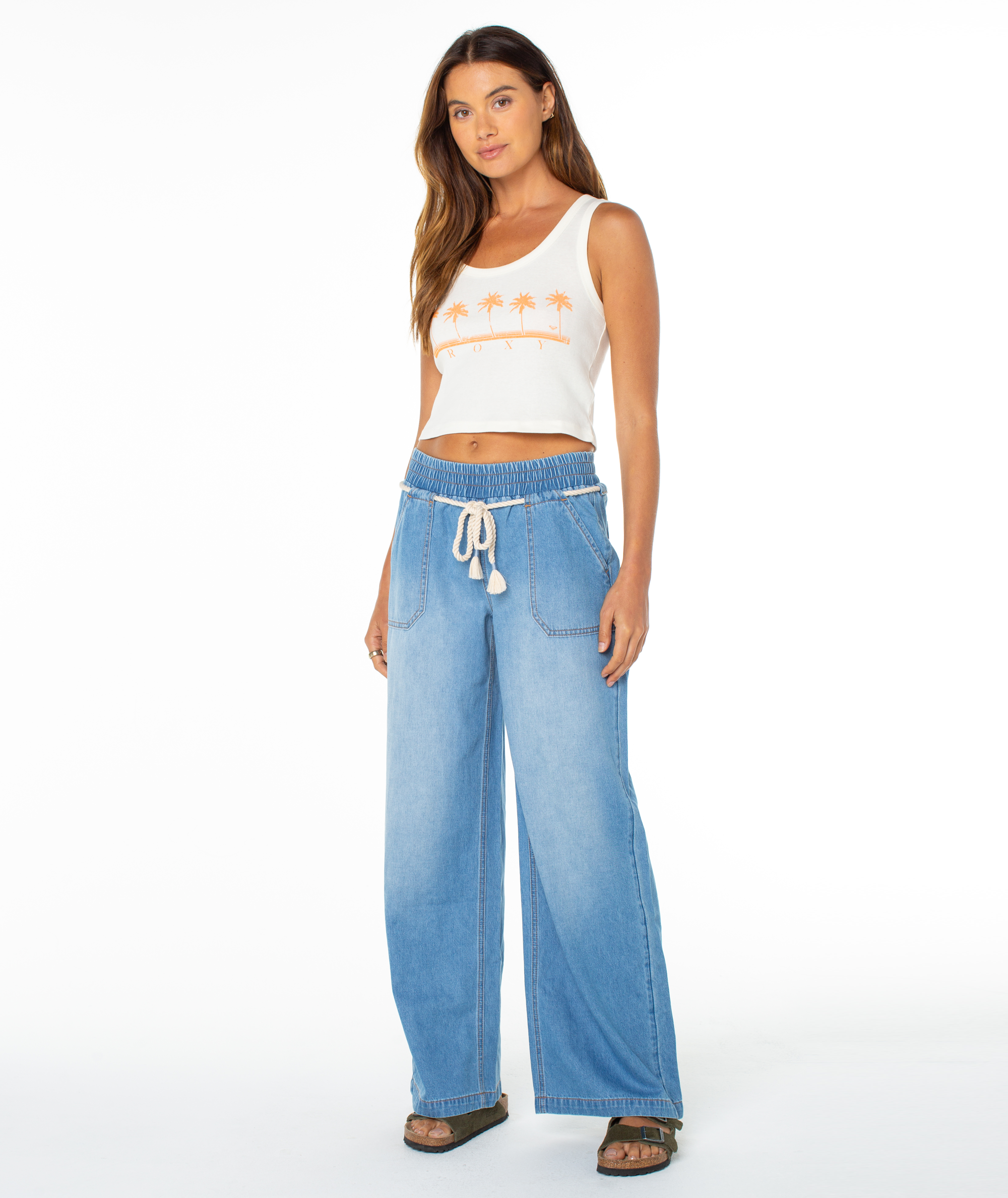 A woman faces forward in Roxy's Five Palm Stripe Tee with an orange palm tree graphic, paired with light blue wide-leg drawstring jeans and dark sandals, showcasing a relaxed coastal vibe against a plain white background.