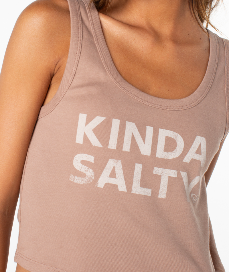 A person models the Roxy Kinda Salty Scoop Baby Rib Tank Top, a sleeveless, light brown cropped tank featuring KINDA SALTY in white on the front. Only their upper torso appears in the image.