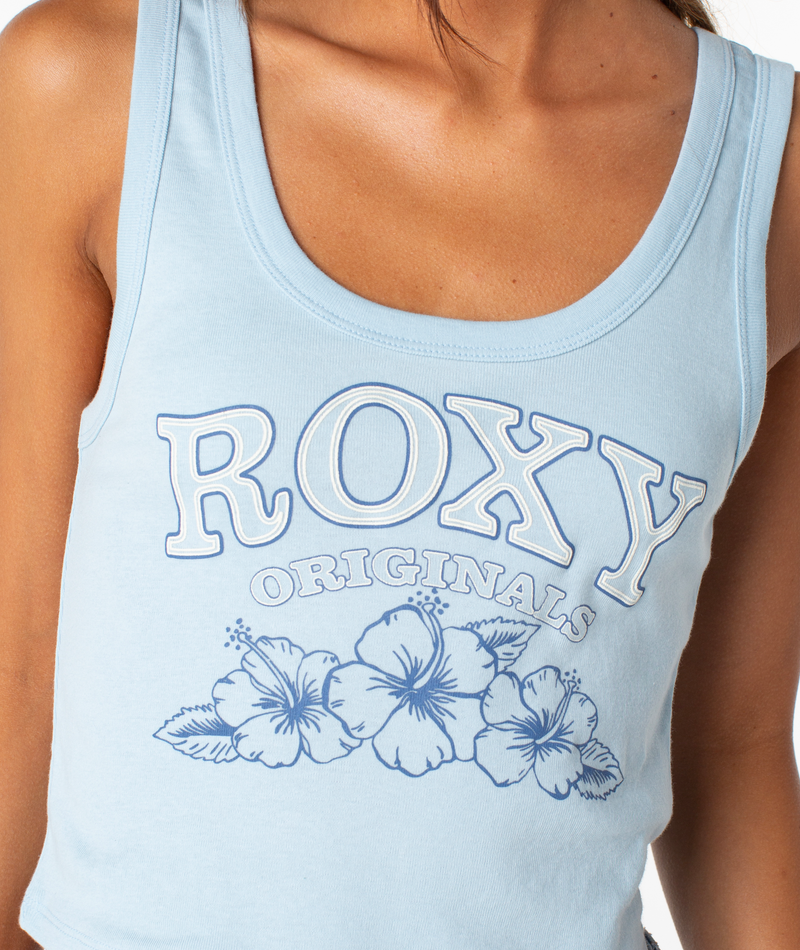 A person wears the Roxy Originals Scoop Baby Rib Tank, a light blue cropped style by Roxy with three hibiscus flowers on the front. Only their upper torso is shown.