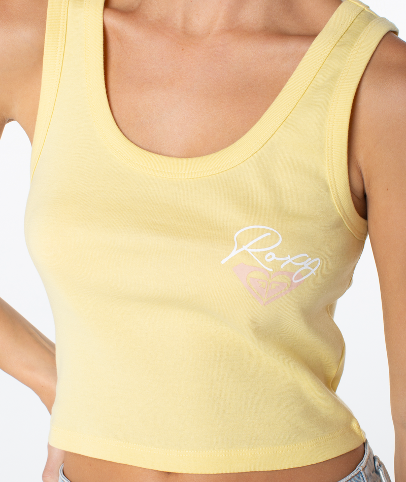 A person wears the Roxy Scoop Baby Rib Tank, a light yellow sleeveless cotton crop top featuring a white and pink heart logo and "Roxy" on the chest, highlighting their bent arm with only their upper body visible.