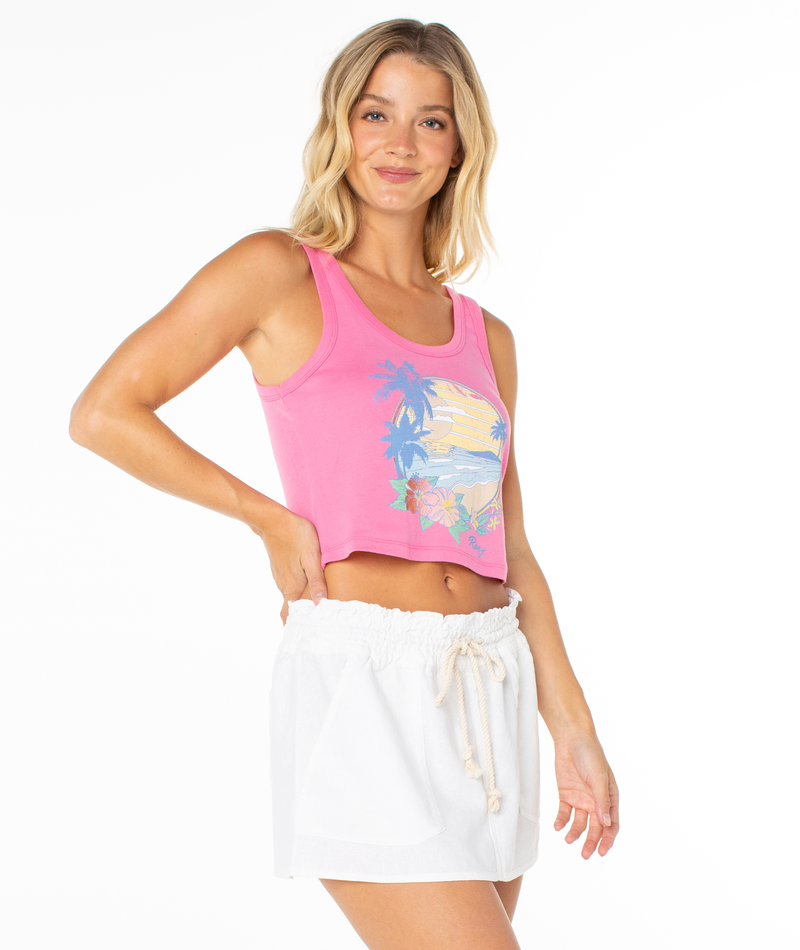 A woman with wavy blonde hair wears the Roxy Scoop Baby Rib Tank, a pink cropped summer essential with a tropical graphic, and white drawstring shorts. She smiles with one hand on her hip against a plain white backdrop.