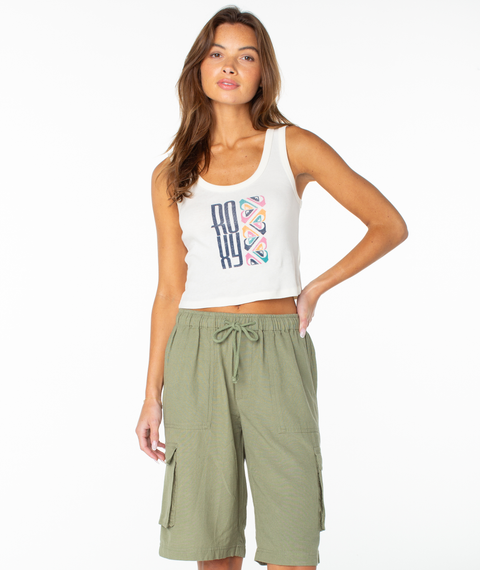 A woman poses against a white background, wearing the Roxy Oceanview Scoop Baby Rib Tank in Cloud Dancer Oceanview, paired with green drawstring cargo shorts. Her long, wavy brown hair falls past her shoulders as her left hand rests on her hip.