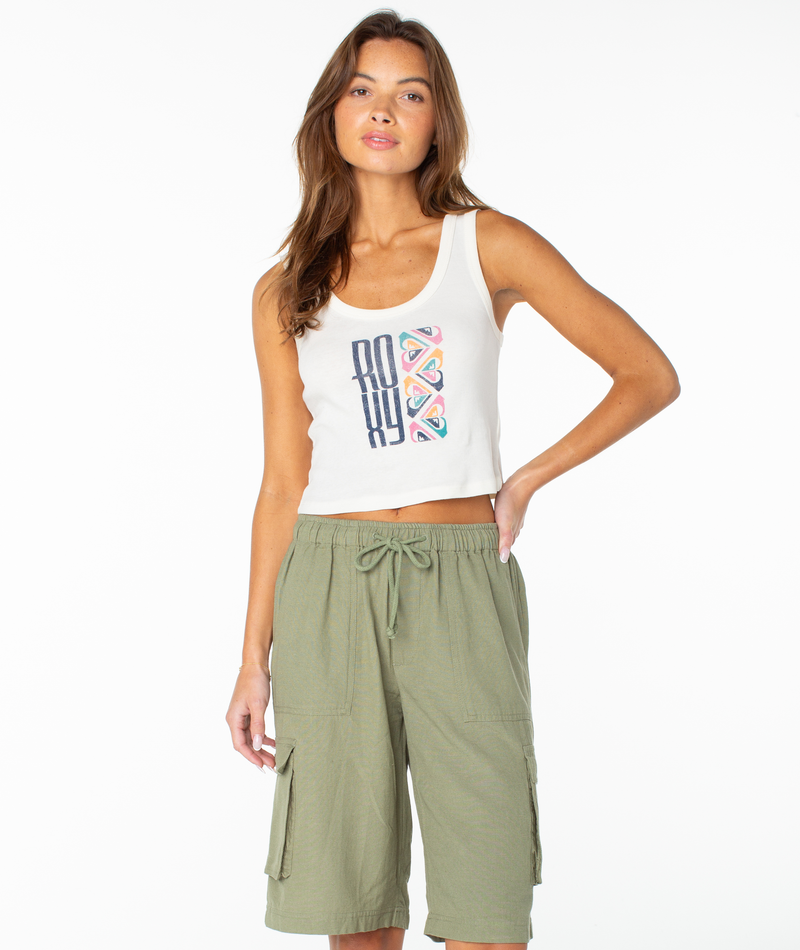 A woman poses against a white background, wearing the Roxy Oceanview Scoop Baby Rib Tank in Cloud Dancer Oceanview, paired with green drawstring cargo shorts. Her long, wavy brown hair falls past her shoulders as her left hand rests on her hip.