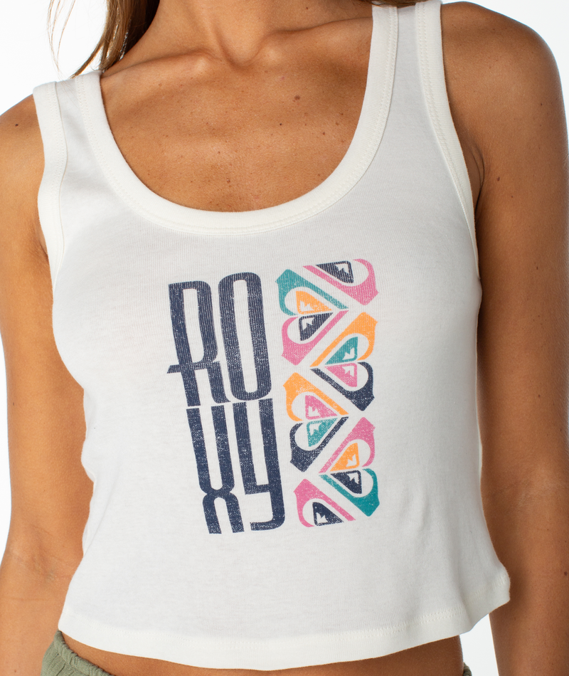 A woman wears the Roxy Oceanview Scoop Baby Rib Tank, a fitted white 90s-inspired top with bold blue "ROXY" lettering and four colorful heart-shaped logos stacked vertically beside the text.