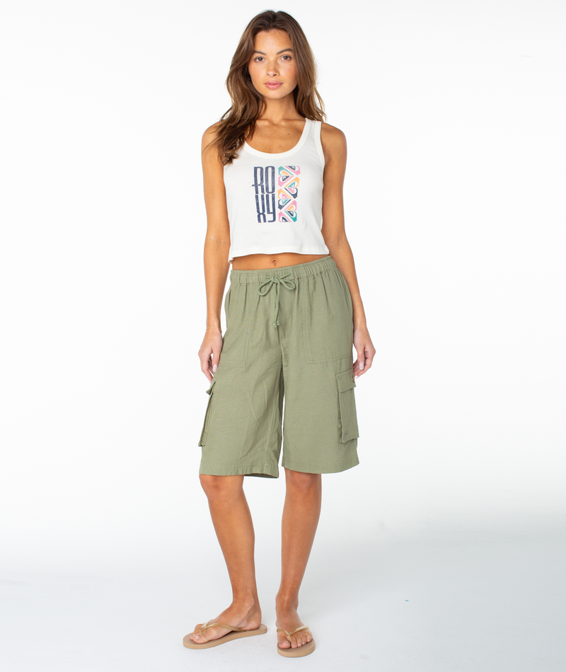 A woman stands against a white background in the Roxy Oceanview Scoop Baby Rib Tank and olive green knee-length cargo shorts, paired with beige flip-flops. Her long hair is loose, completing her retro beach-inspired look.