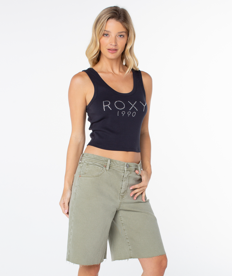 A woman poses in the Roxy Shine Bright Scoop Baby Rib Tank, a sleeveless black crop top featuring “ROXY 1990” text, paired with light green knee-length shorts against a plain white background, channeling a retro-inspired vibe reminiscent of the classic ‘90s baby tee.