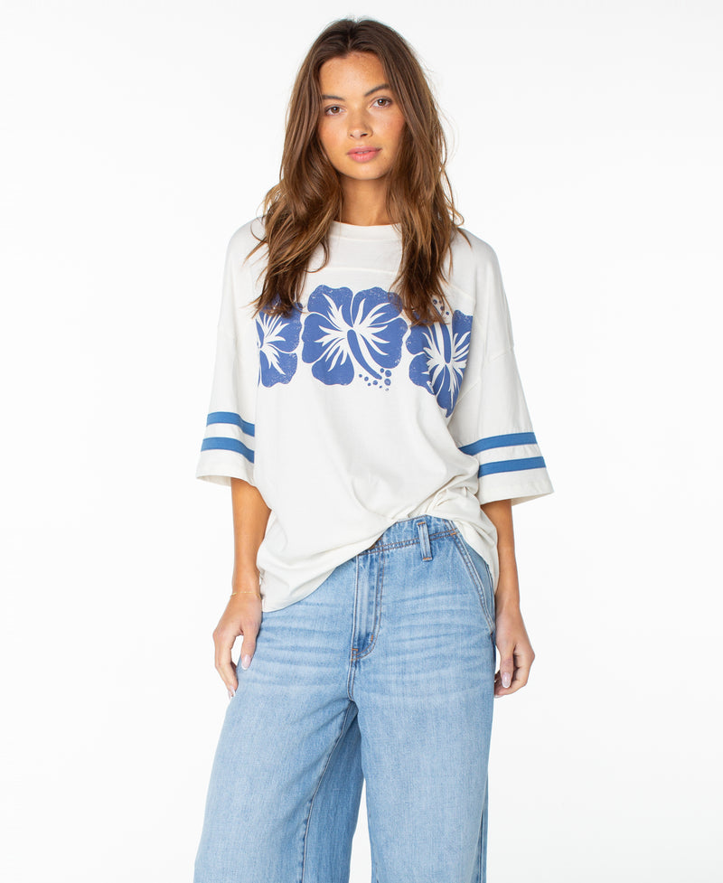 A woman with long brown hair wears the Roxy Game Day Tee in Cloud Dancer Flower Block—a loose, boxy-fit white tee featuring blue hibiscus flowers, retro accents, and blue striped sleeves. She pairs it effortlessly with light blue wide-leg jeans and stands against a plain white background.
