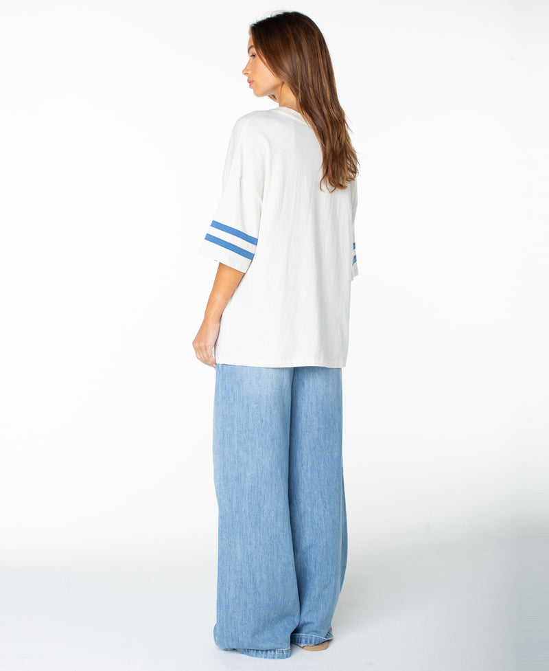 A woman stands facing away against a plain white background, wearing the Roxy Game Day Tee—an easy-fitting white shirt featuring retro blue sleeve stripes—paired with wide-leg light blue jeans.