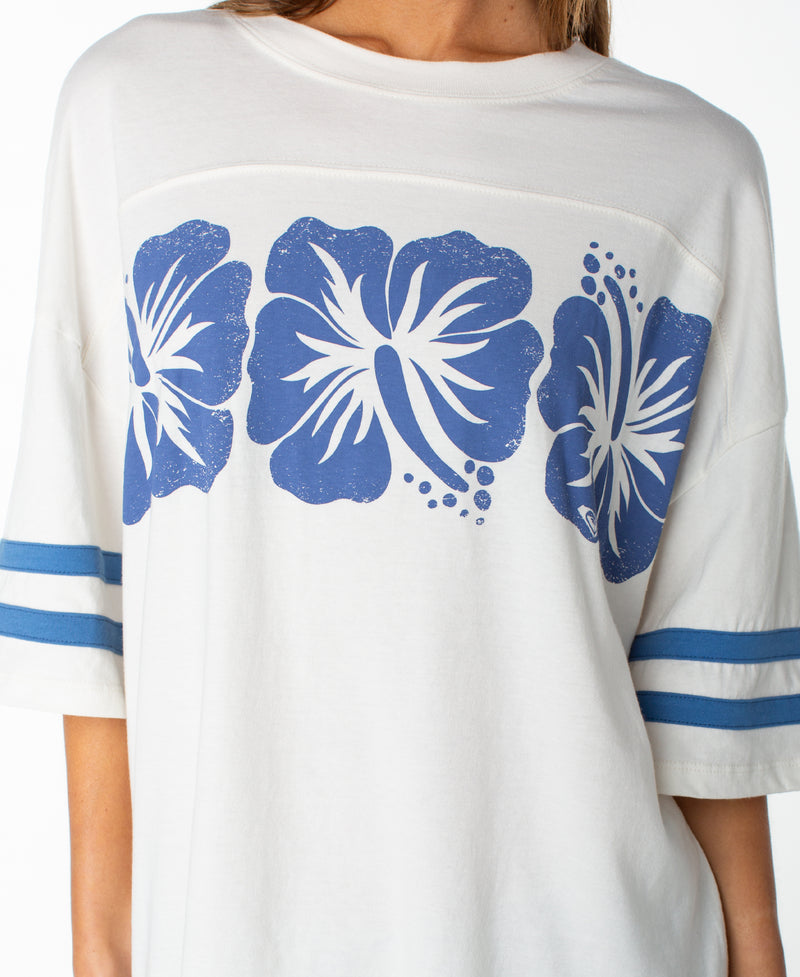 A person wearing the Roxy Game Day Tee, featuring a boxy fit, blue sleeve stripes, and three bold blue hibiscus flowers printed across the chest.