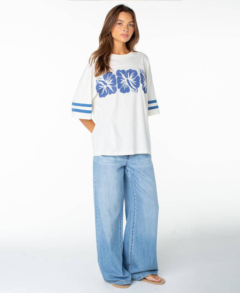 A woman stands against a white background wearing the Roxy Game Day Tee—a boxy fit white shirt adorned with blue hibiscus flowers and blue striped sleeves, featuring subtle retro details—paired with light blue wide-leg jeans and sandals.