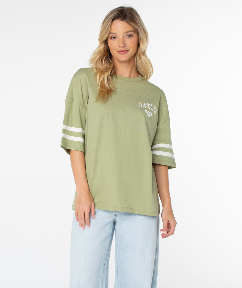 A woman with long blonde hair wears the Roxy Game Day Tee in Oil Green Classic Uni—a loose, boxy-fit top with white retro stripes on the sleeves and the ROXY logo—paired with light blue jeans and standing against a plain white background.