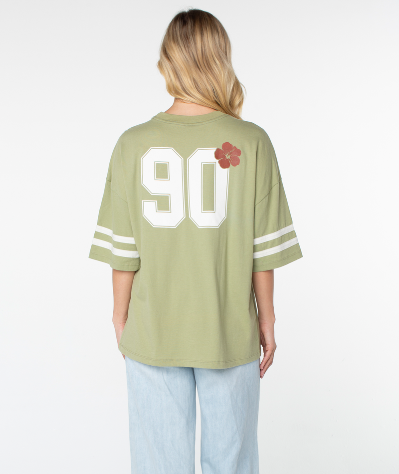 A woman with blonde hair is seen from the back wearing the Roxy Game Day Tee in light green, which features white striped sleeves, retro-inspired details including the number 90, and a red flower graphic on the back. The boxy fit of her tee stands out as she pairs it with light blue jeans.