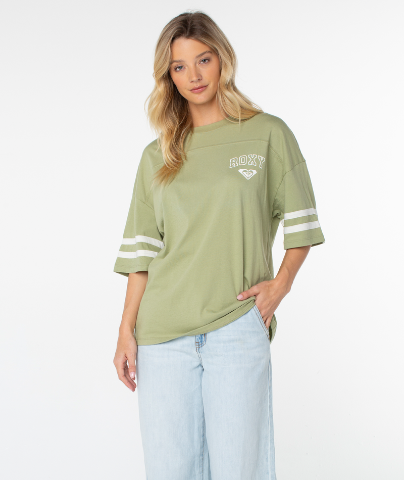 A woman stands against a plain white background with one hand in her pocket, wearing the Roxy Game Day Tee—a loose, light green top featuring retro-inspired white sleeve stripes and a relaxed boxy fit—paired with light blue jeans.