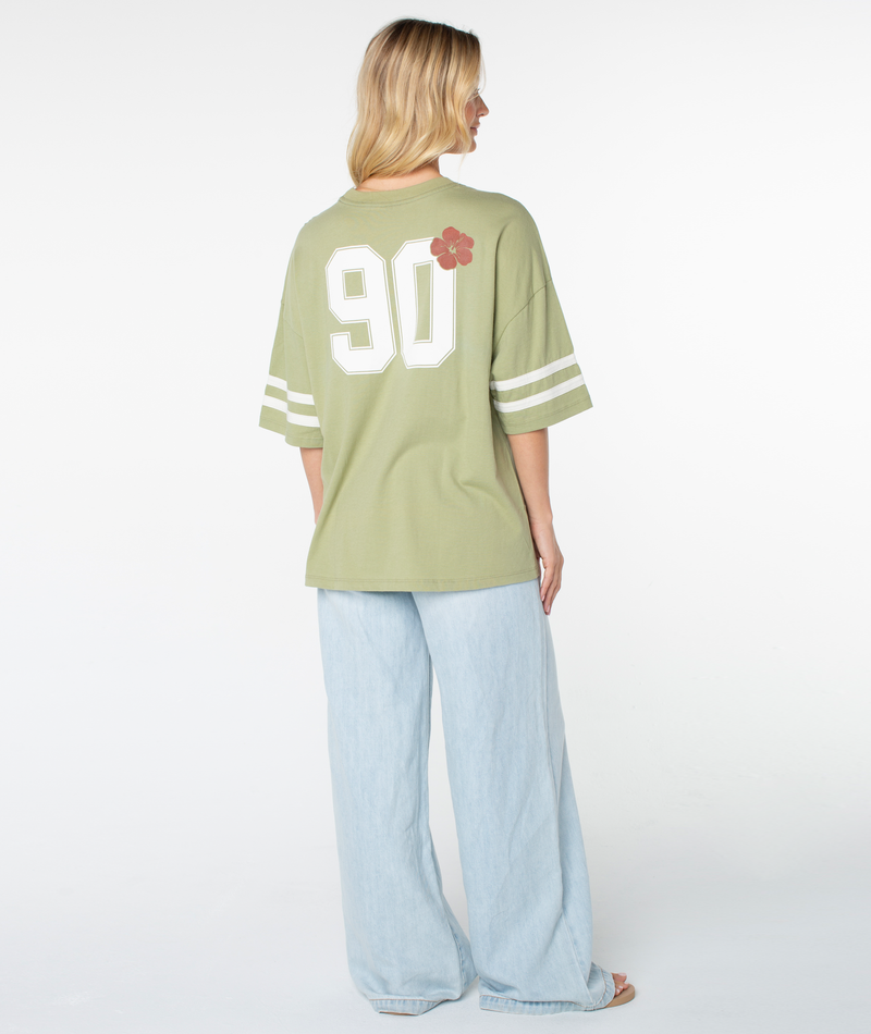 A person stands facing away, wearing the Roxy Game Day Tee in sage green, which features a boxy fit, white sleeve stripes, the number 90, and a red flower graphic on the back. The tee is paired with light blue wide-leg pants that highlight its retro details.