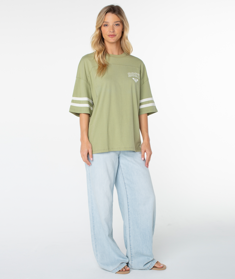 A woman with long blonde hair models the Roxy Game Day Tee—an oversized, boxy-fit light green top featuring retro white sleeve stripes—paired with light blue wide-leg jeans, standing in front of a plain white background.