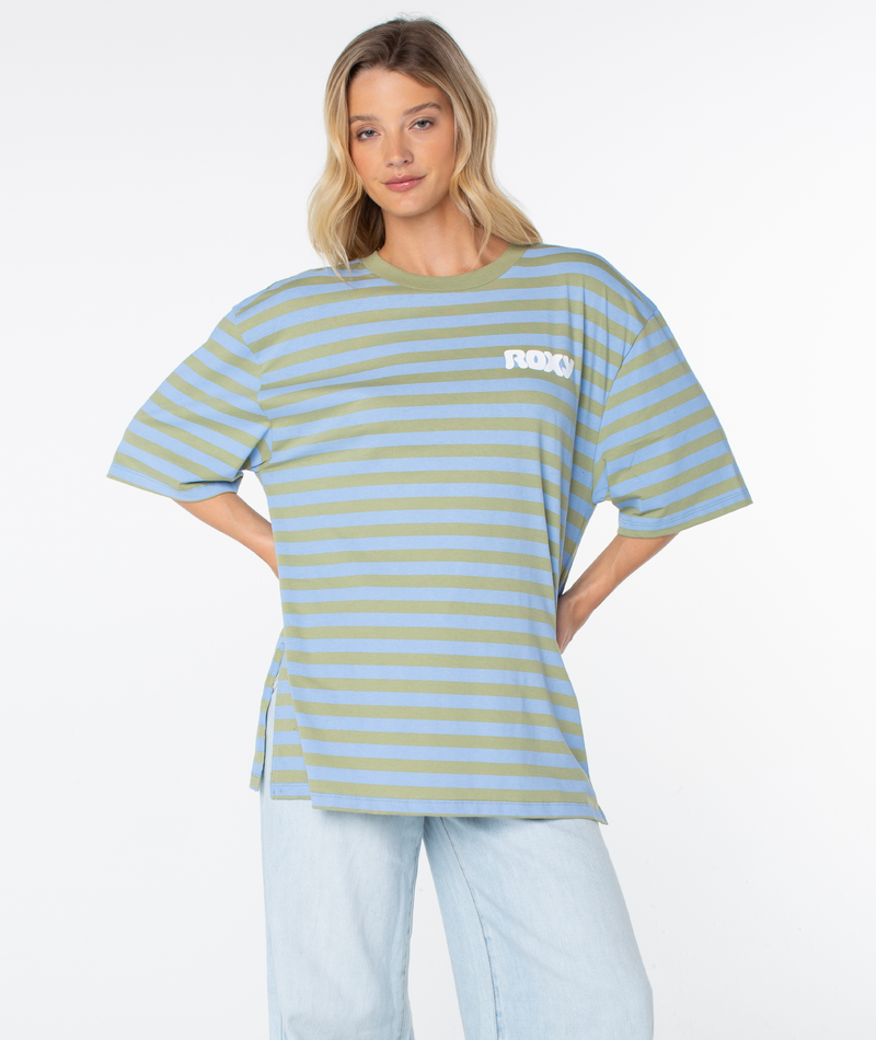 A woman stands with her hands on her hips against a plain white background, wearing light blue jeans and the Roxy Old School Tee in Oil Green Wave Riders Club. This warm-weather staple features an oversized silhouette for effortless comfort and laid-back style, with “ROXY” written on the chest.