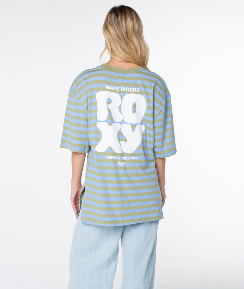 A person with long blonde hair faces away, wearing the Roxy Old School Tee—an oversized, loose-fit striped green and blue shirt with “WAVE RIDERS ROXY SURFING SINCE 1990” printed on the back—styled as a warm-weather staple and paired with light blue wide-leg pants.