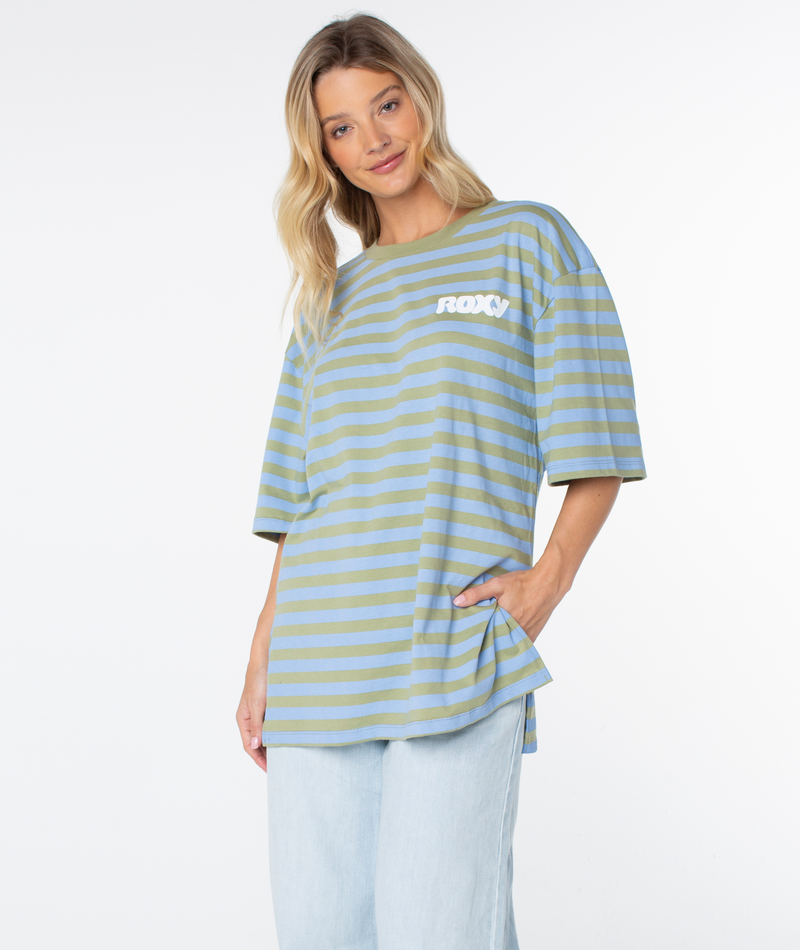 A woman is wearing the Roxy Old School Tee—a loose, striped blue and green T-shirt with bold ROXY lettering on the chest—paired with light blue jeans. She poses with one hand in her pocket against a plain white background, showcasing the tee’s oversized silhouette, perfect as a warm-weather staple.