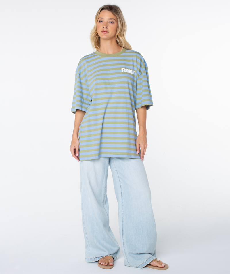 A woman stands against a plain white background wearing the Roxy Old School Tee—an oversized tee with loose green and blue stripes and bold FROG text—paired with light wide-leg jeans and sandals, embodying an effortless warm-weather look.