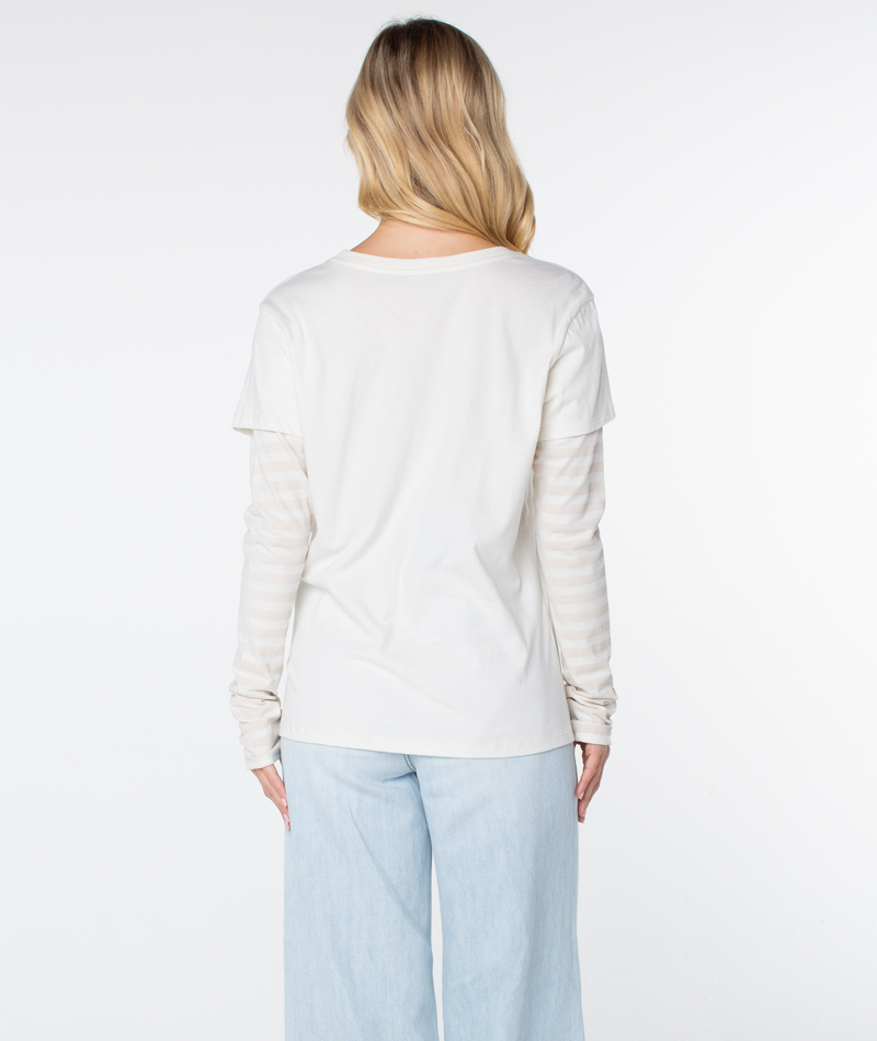 A person with long blonde hair stands facing away, wearing the Vintage Roxy Two-fer Top by Roxy layered over a striped shirt and paired with light blue pants against a white background.