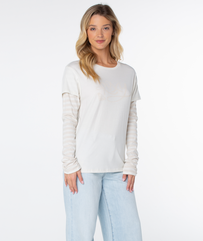 Woman with long blonde hair models the Vintage Roxy Two-fer Top by Roxy— a 90’s-inspired white tee layered over a striped long sleeve, paired with light blue jeans and a gentle smile against a white background.