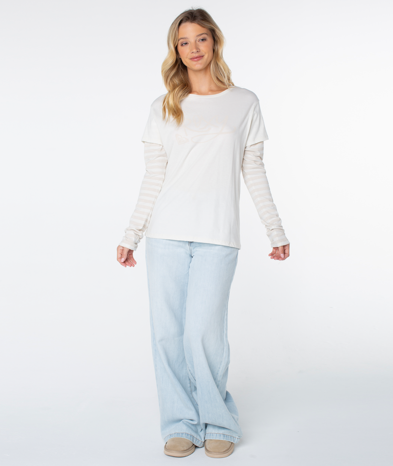 A woman with long blonde hair wears the Vintage Roxy Two-fer Top in Cloud Dancer Vintage Roxy by Roxy, light blue jeans, and beige shoes, standing against a white background.