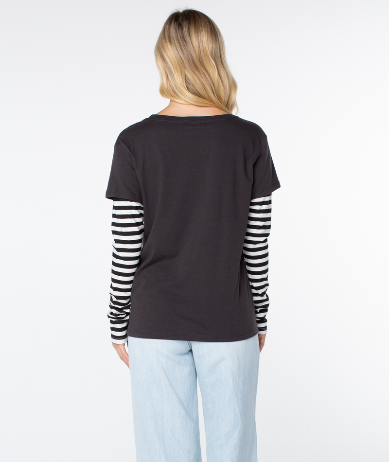 A person with long blonde hair faces away, showcasing a modern coastal look in the Roxy Kinda Salty Two-fer Top and light blue jeans against a plain white background.